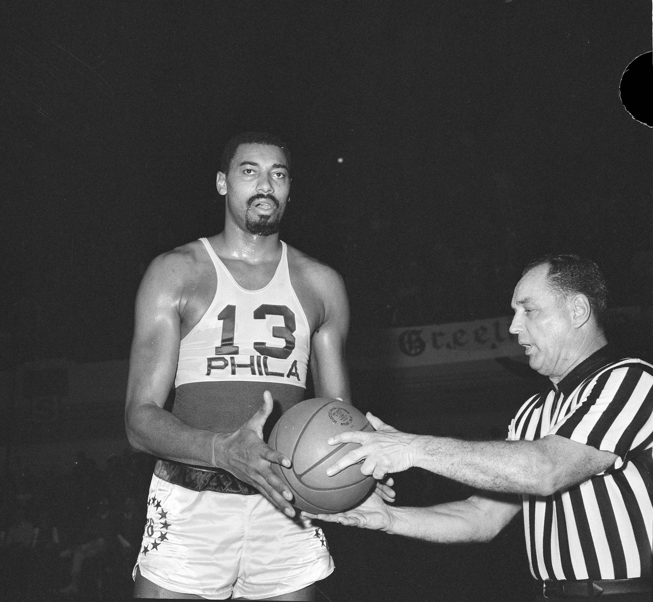 Wilt Chamberlain of the Philadelphia 76ers, who became the second player in the history of the NBA to score 20,000 points, accepts the ball from referee Willie Smith as game with New York Knicks was halted in Philadelphia, Jan. 2, 1965. Presentation was made after Chamberlain sank foul shot for 20,000th point. Chamberlain hopes to surpass the all-time point scoring record of 20,880, held by Bob Pettit, St. Louis Hawks star, before the season ends. 76ers won the game, 133-122. (AP Photo/Bill Ingraham)